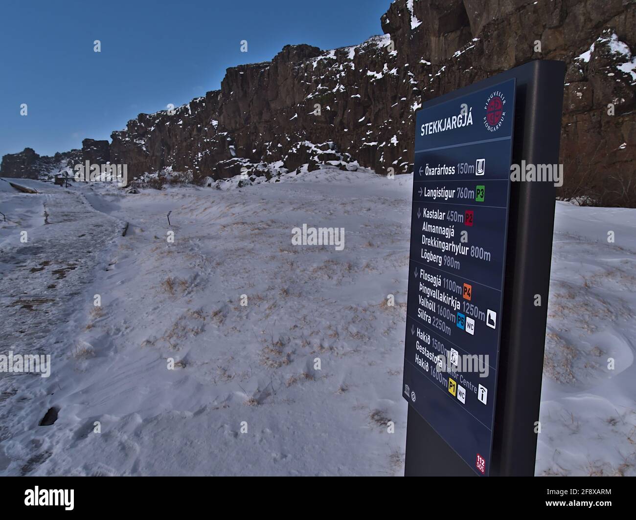 Vista del cartello con destinazioni e distanze accanto al sentiero innevato nel Parco Nazionale di Thingvellir nella stagione invernale con rocce vulcaniche. Foto Stock
