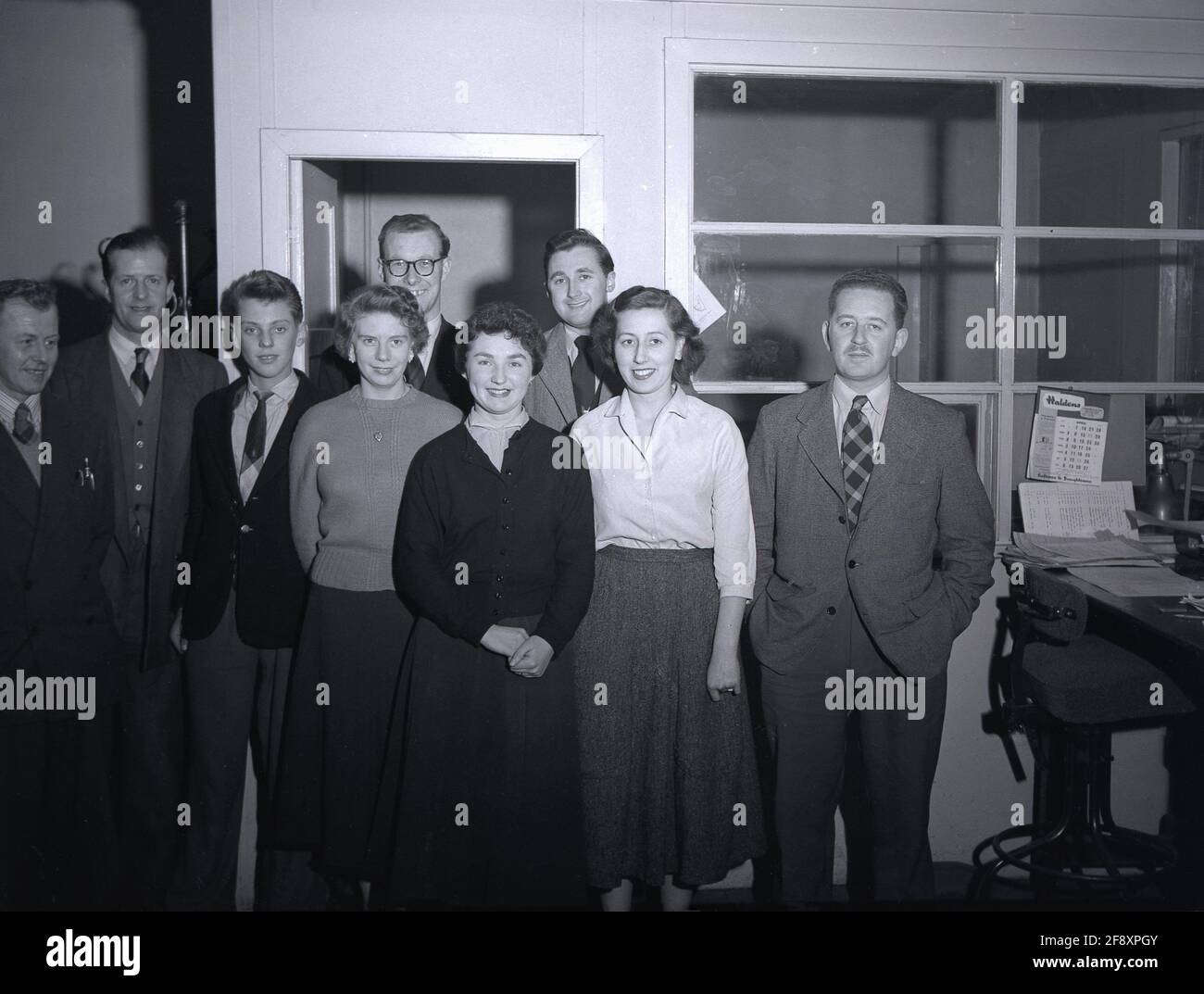 Anni '50, storico, all'interno di un ufficio aziendale, un gruppo di uomini e donne di amministrazione si levano in piedi insieme per una foto, Inghilterra, Regno Unito. Il luogo di lavoro in ufficio in questa era era formale e i dipendenti si aspettavano di vestirsi in un certo modo. Sia per gli uomini che per le donne, un abito di lana su misura intelligente, ha presentato un'immagine professionale e sarebbe stato l'abbigliamento appropriato in un ambiente aziendale ufficio al thh time. Come si può vedere, anche il giovane ragazzo, l'ufficio junior, indossa una giacca e cravatta. Foto Stock