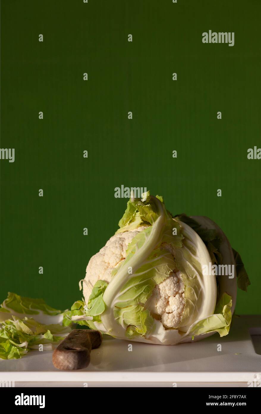 Un cavolfiore intero su un tagliere bianco con coltello e uno sfondo colorato a strisce verdi Foto Stock
