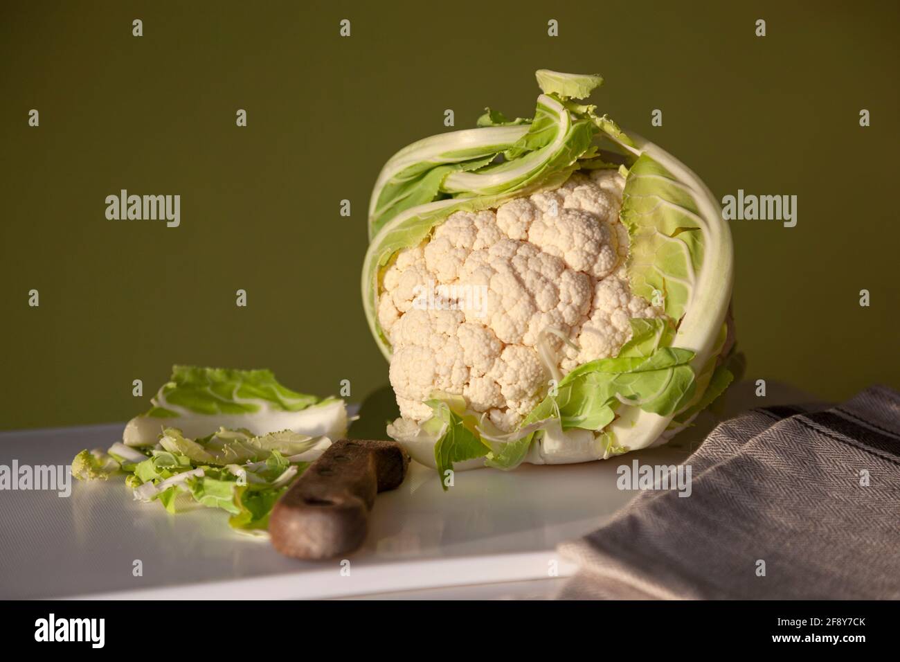 Un cavolfiore intero su un tagliere bianco con coltello, tovagliolo di lino e uno sfondo di colore verde chiaro. Foto Stock