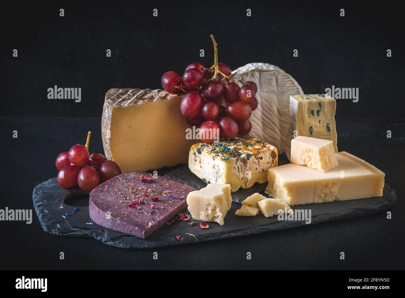 Varietà di formaggio esclusivo e colorato, decorato con uve rosse, su sfondo nero Foto Stock