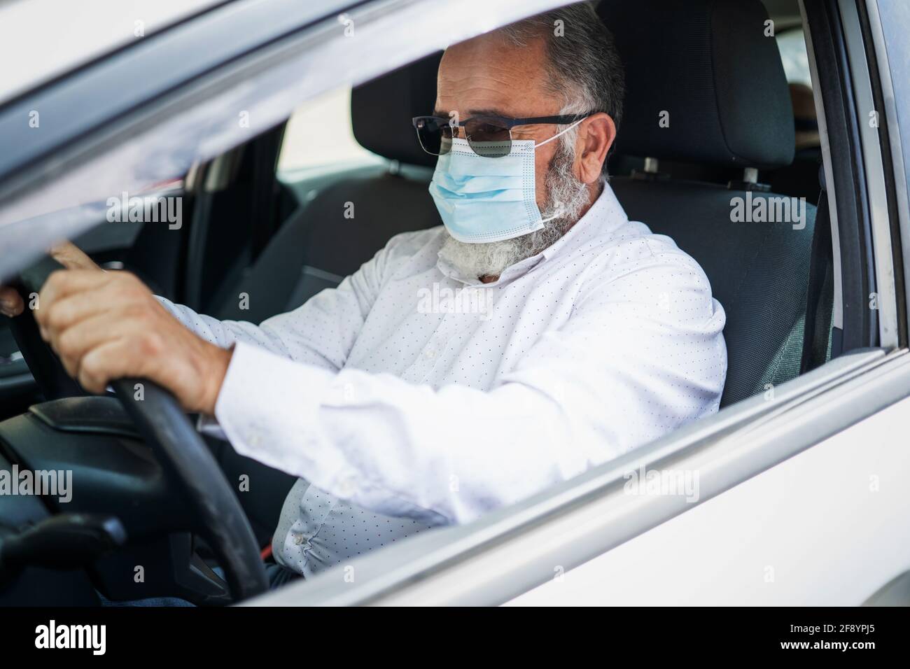 Un uomo anziano in una maschera medica chirurgica che guida un'automobile. Concetto pandemico di coronavirus. Foto Stock
