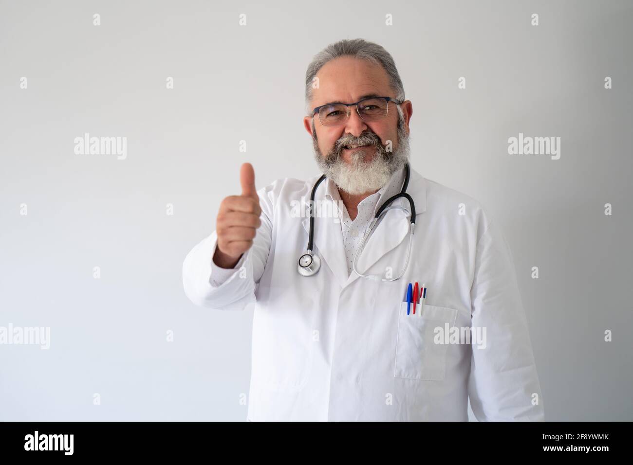 Ritratto di dottore anziano in camice bianco sorridente con pollici su sfondo bianco. Foto Stock