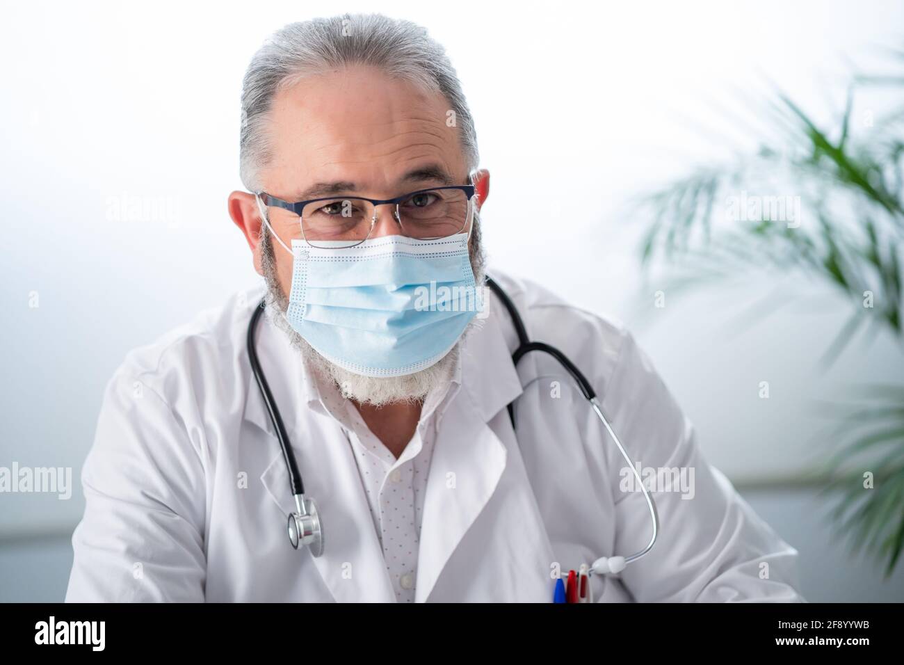 Ritratto di dottore anziano in camice bianco e maschera facciale che guarda la macchina fotografica nel suo ufficio. Foto Stock