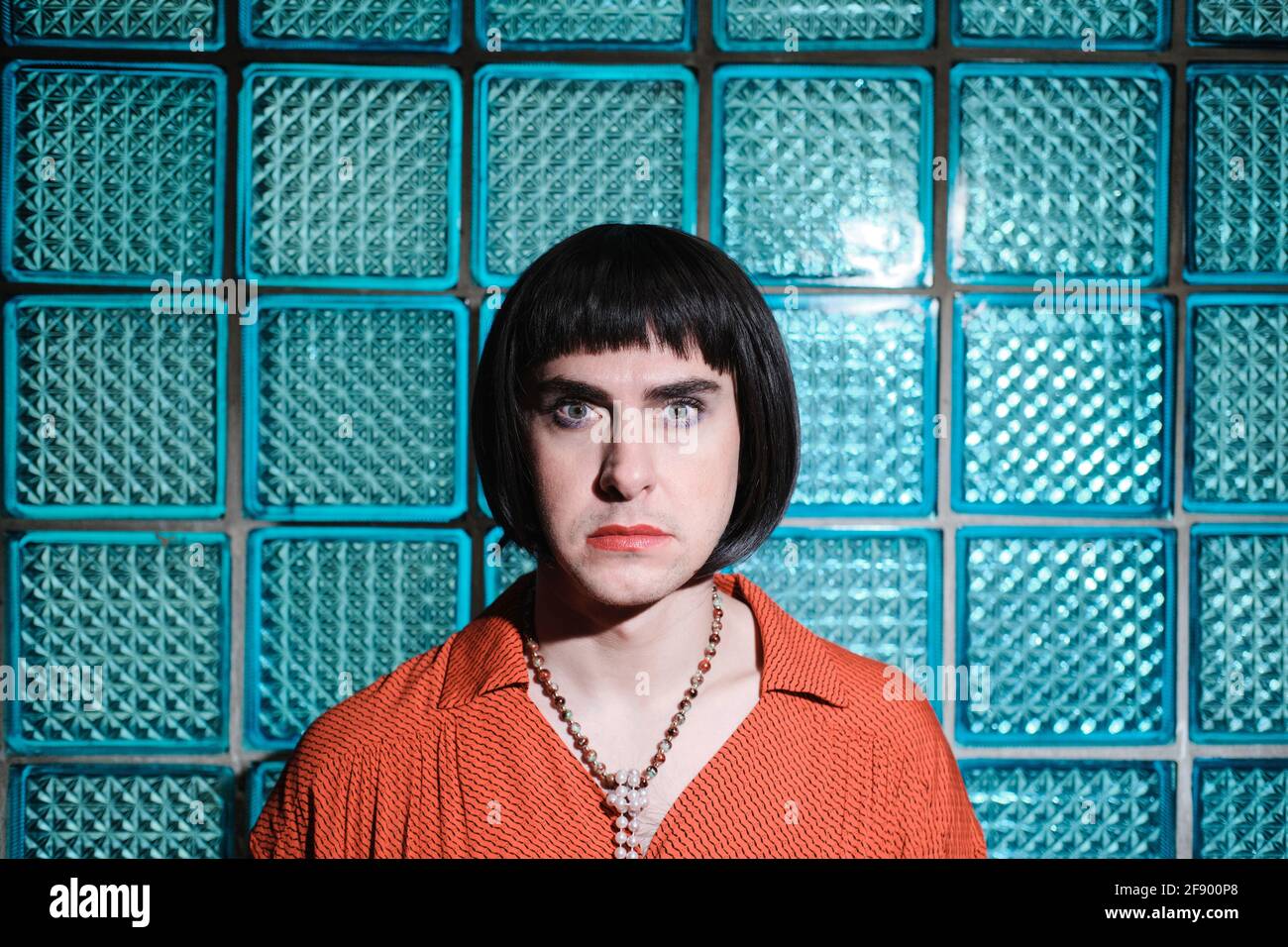 Ritratto di transessuale in parrucca nera corta guardando la macchina fotografica in piedi contro la parete di vetro blu Foto Stock