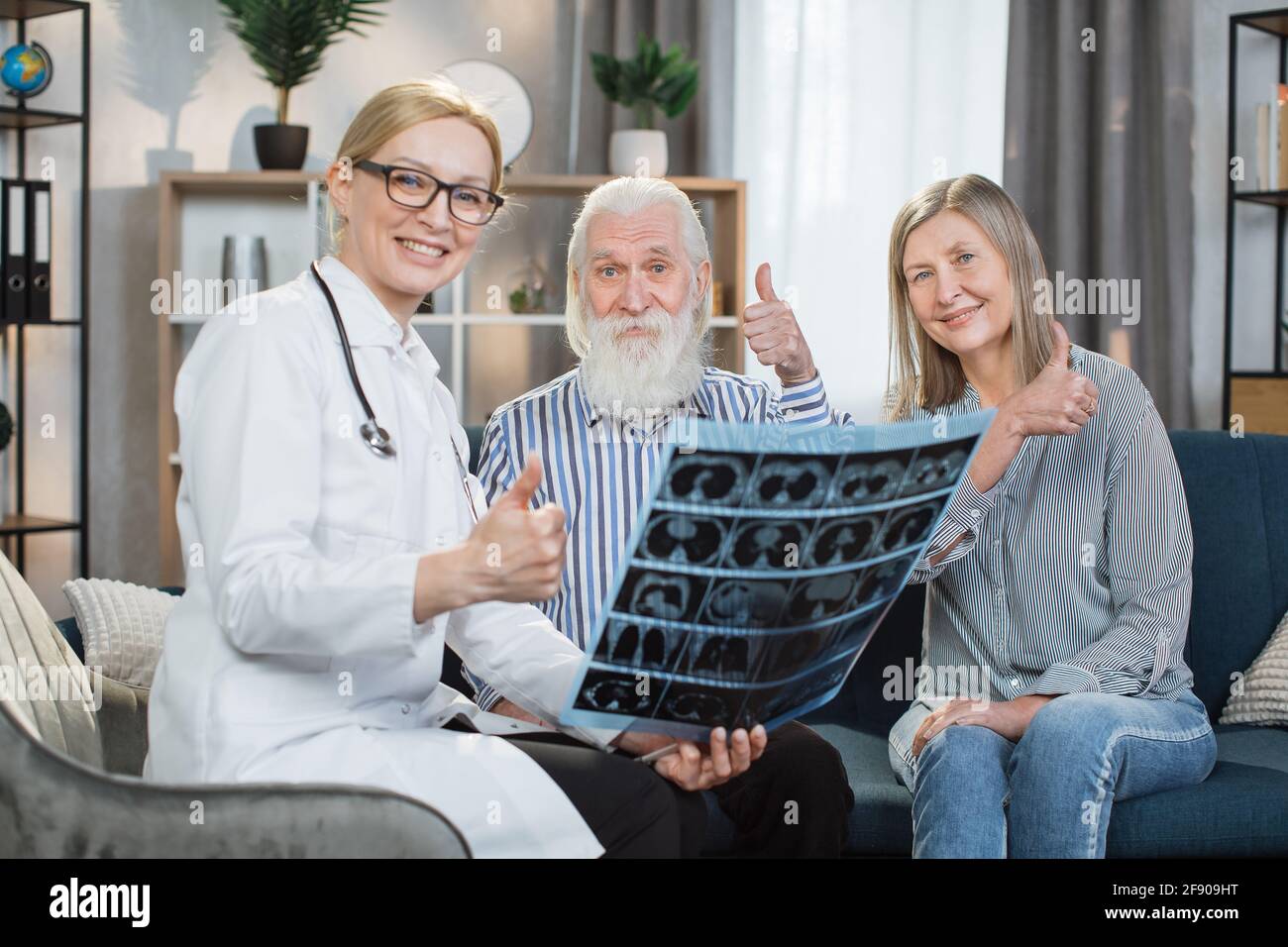Buone notizie, salute forte. Bella donna sorridente dottore soddisfatto dei risultati della scansione a raggi X dei suoi pazienti, felice uomo anziano sopportato e donna piacevole, mostrando pollici fino alla macchina fotografica. Foto Stock