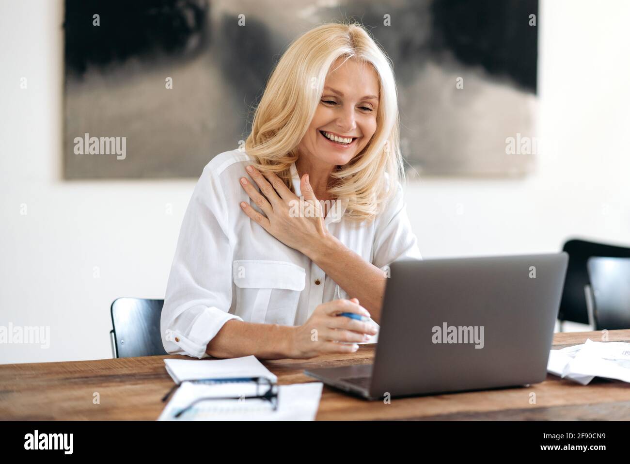 Amichevole, sicuro elegante bionda matura, donna di affari o manager, usa il laptop, chattando in videoconferenza con i colleghi o il cliente, gesturing con la mano e sorridente. Video meeting online Foto Stock
