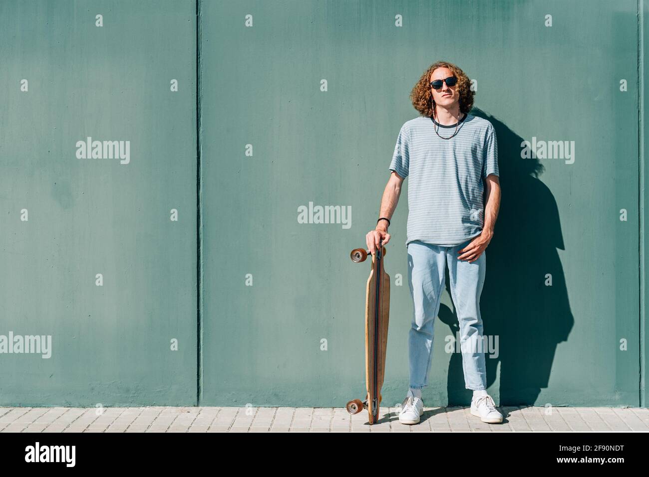 ritratto orizzontale di un giovane skateboarder caucasico che guarda la fotocamera. Indossa abiti e occhiali da sole casual. Sfondo verde Foto Stock
