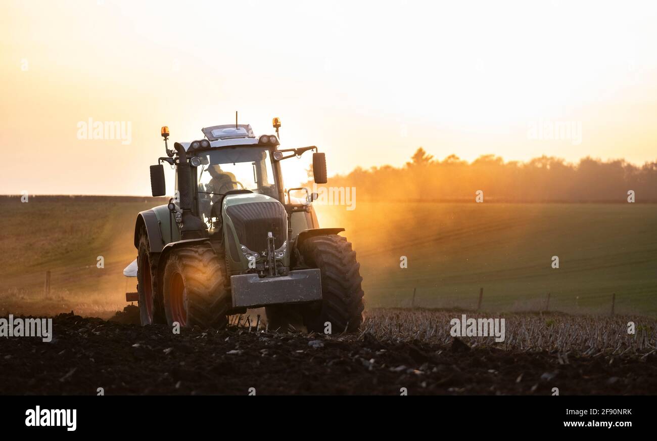 Aratura di un campo al tramonto con un trattore e un aratro, pronti per i raccolti in un'azienda agricola Foto Stock