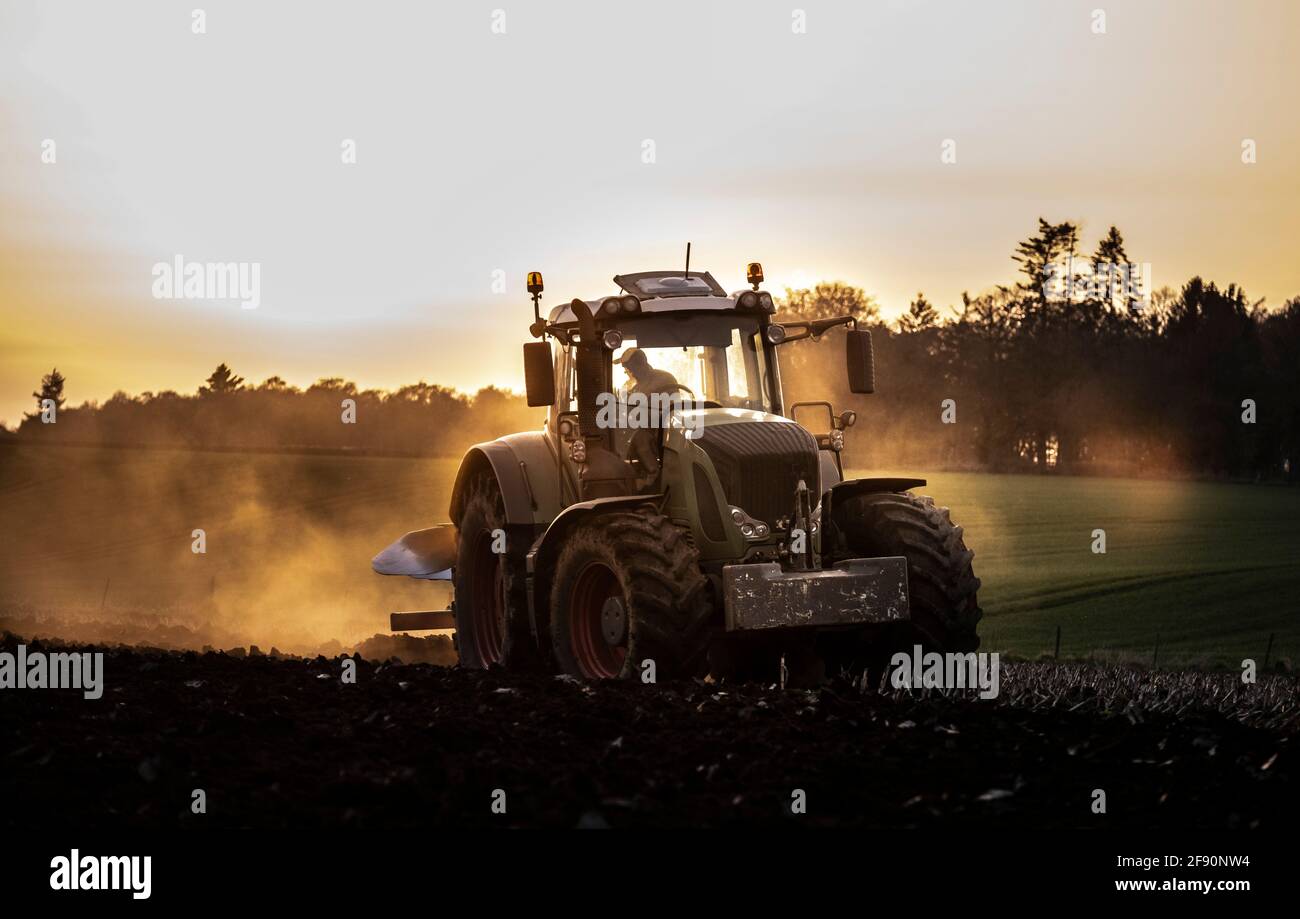 Aratura di un campo al tramonto con un trattore e un aratro, pronti per i raccolti in un'azienda agricola Foto Stock