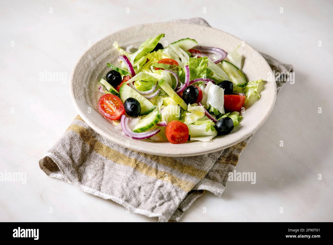 Insalata di verdure classiche con pomodori, cetrioli, cipolle, foglie di insalata e olive nere in piastra bianca in ceramica su tovagliolo di stoffa. Sfondo in marmo bianco. Foto Stock