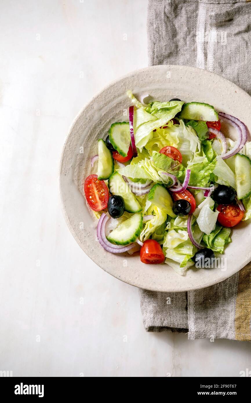 Insalata di verdure classiche con pomodori, cetrioli, cipolle, foglie di insalata e olive nere in piastra bianca in ceramica su tovagliolo di stoffa. Sfondo in marmo bianco. Foto Stock