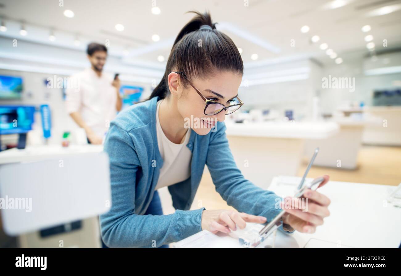 Giovane soddisfatto elegante affascinante bella ragazza che prova il nuovo modello di un mobile dalla scrivania nel negozio di tecnologia. Foto Stock