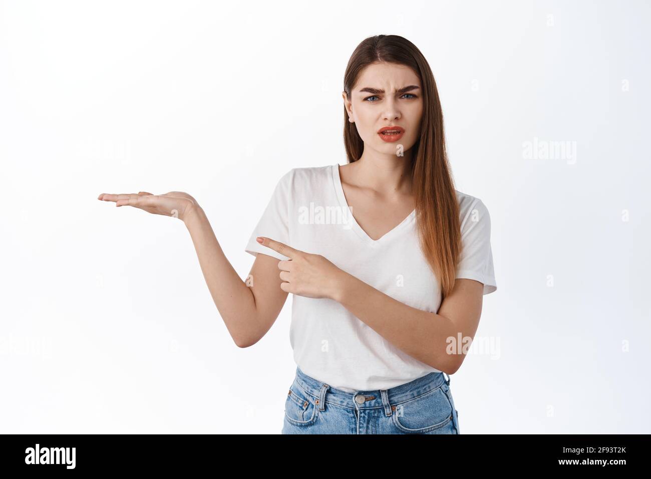Donna confusa che tiene qualcosa di strano in mano aperta, puntando al palmo con il tuo logo prodotto contro lo spazio di copia, interrogato su articolo, bianco Foto Stock