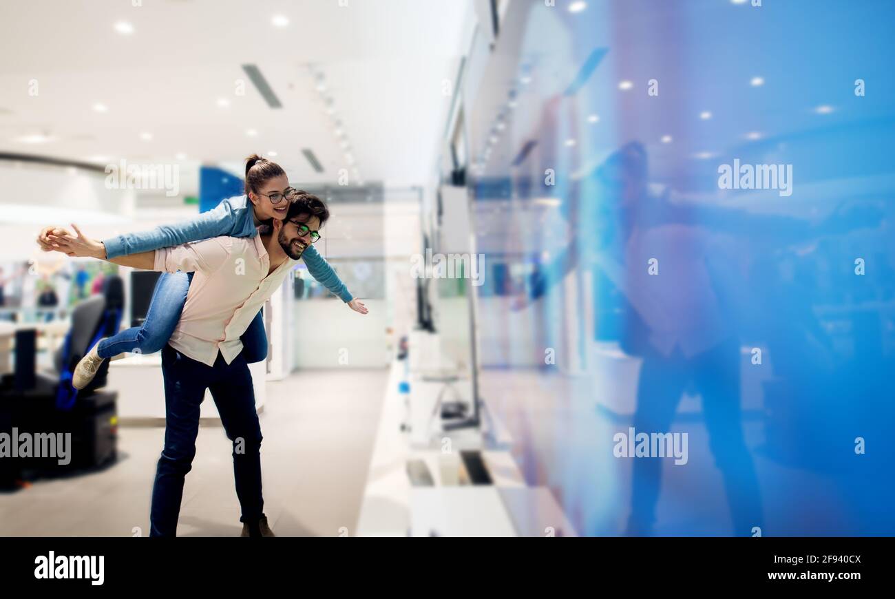 Bella giovane coppia moderna divertirsi mentre si fa shopping nel negozio di elettronica con un giro in piggyback e a braccia aperte. Foto Stock