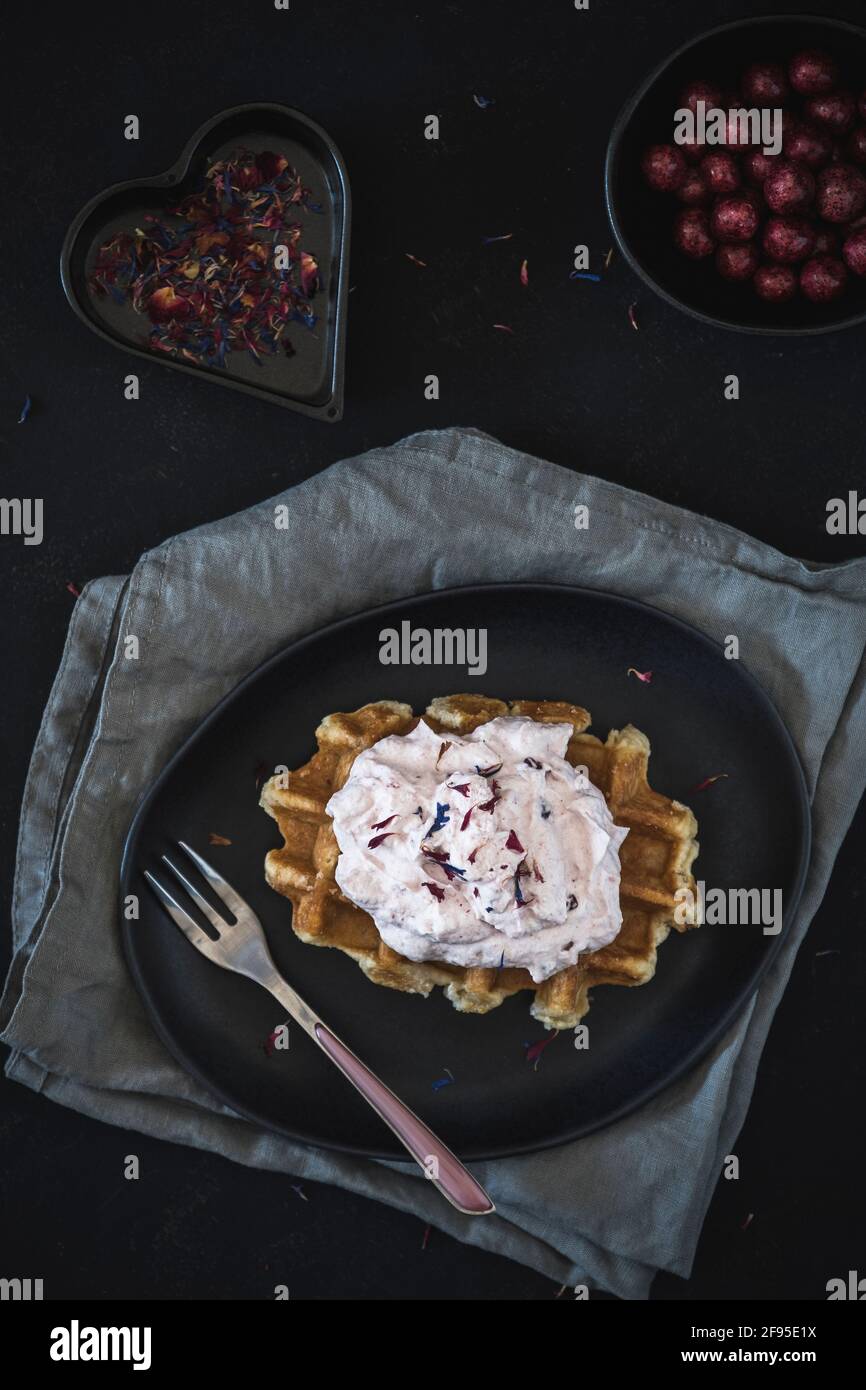 Waffle belgi con crema di mirtilli rossi, adagiati su un piatto nero e tovaglioli grigi, decorati con forchetta di rose e petali colorati e agili Foto Stock