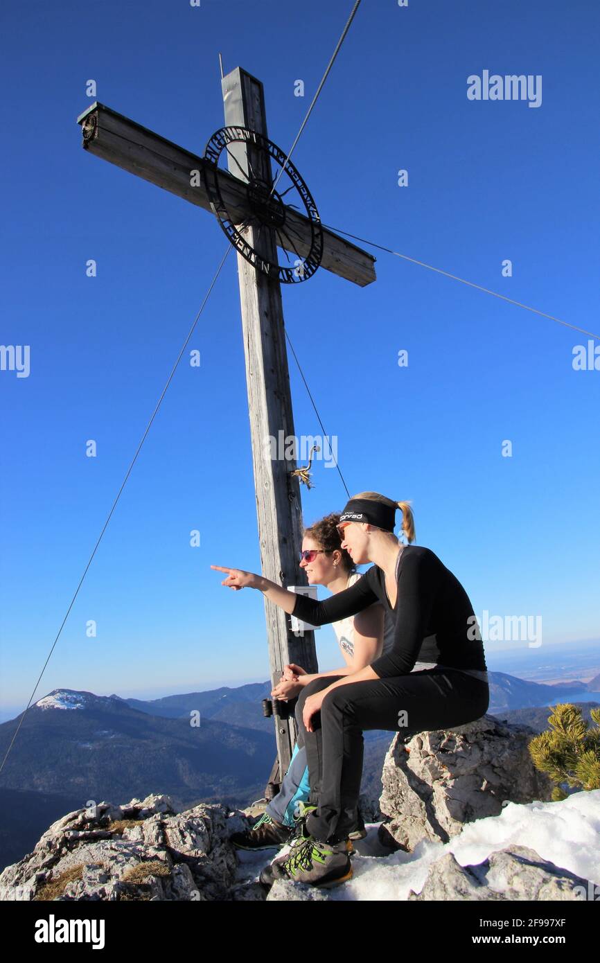 Escursione invernale a Signalkopf (1895 metri) 2 giovani donne si siedono alla croce, 1 donna punta con il dito al meraviglioso panorama montano, Europa, Germania, Baviera, alta Baviera, Isar Valley, Krün Foto Stock