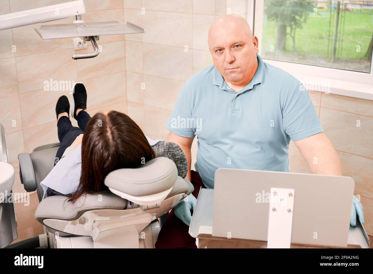 Dentista che guarda la fotocamera mentre si siede accanto alla paziente. Donna sdraiata in sedia dentale durante il trattamento stomatologico in centro medico. Concetto di odontoiatria, stomatologia e cura dentale Foto Stock