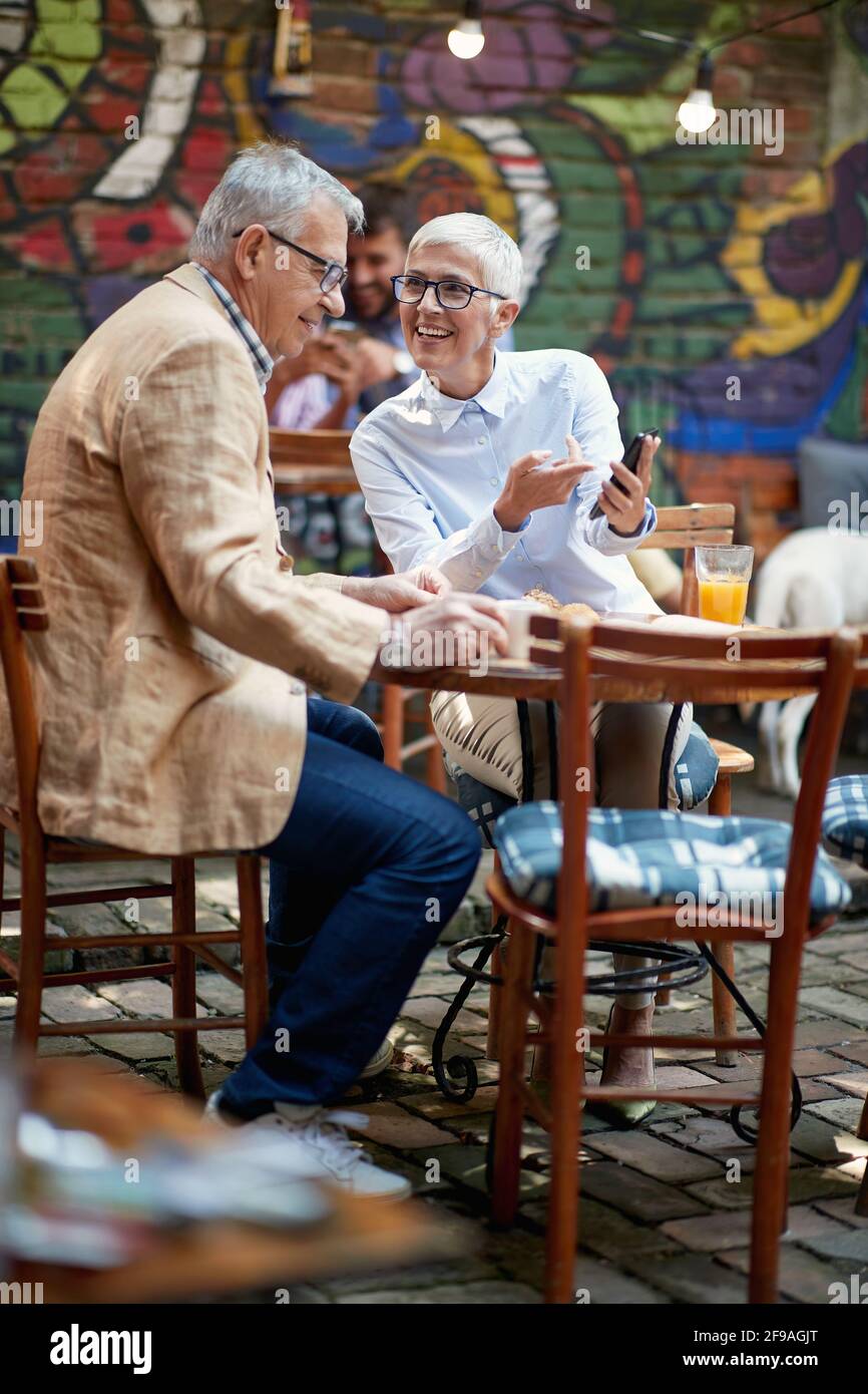 Una coppia più anziana guarda i contenuti di uno smartphone mentre sorseggia un drink in un'atmosfera rilassata al bar. Tempo libero, bar, amicizia, all'aperto Foto Stock