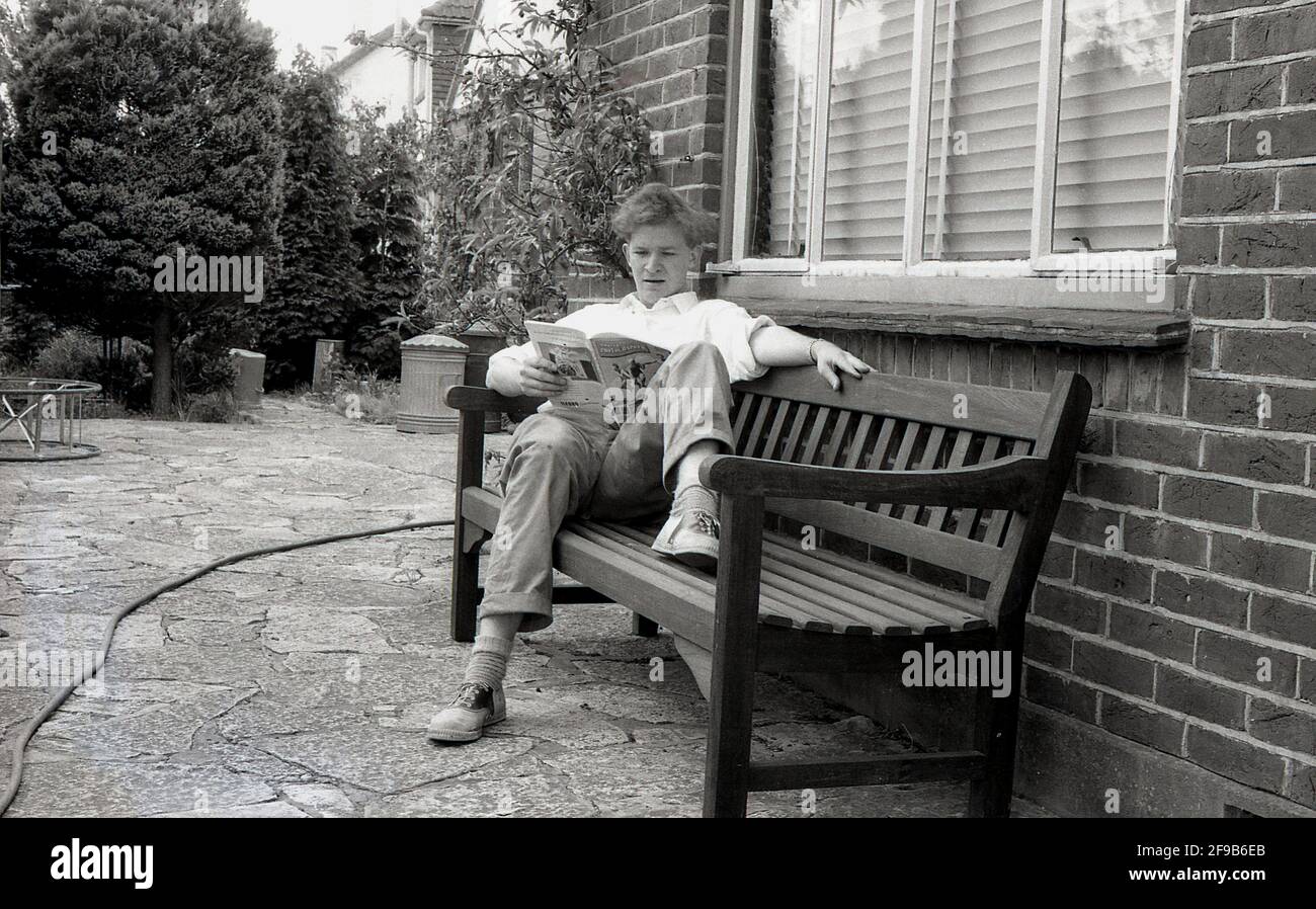 Anni '60, storico, su un patio lastricato, al di fuori di una casa, un giovane seduto su una panchina di legno leggendo la rivista, 'Amateur Photographer', Inghilterra, UK. La popolare rivista di fotografia britannica - la più antica rivista fotografica settimanale al mondo per i consumatori - è stata pubblicata per la prima volta nell'ottobre 1884 ed è ancora in fase di pubblicazione oggi. Foto Stock