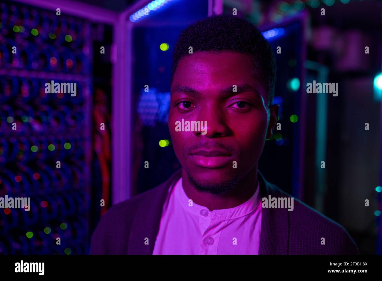 Ritratto di un giovane e serio ingegnere di data center afroamericano in piedi armadio server in camera oscura Foto Stock