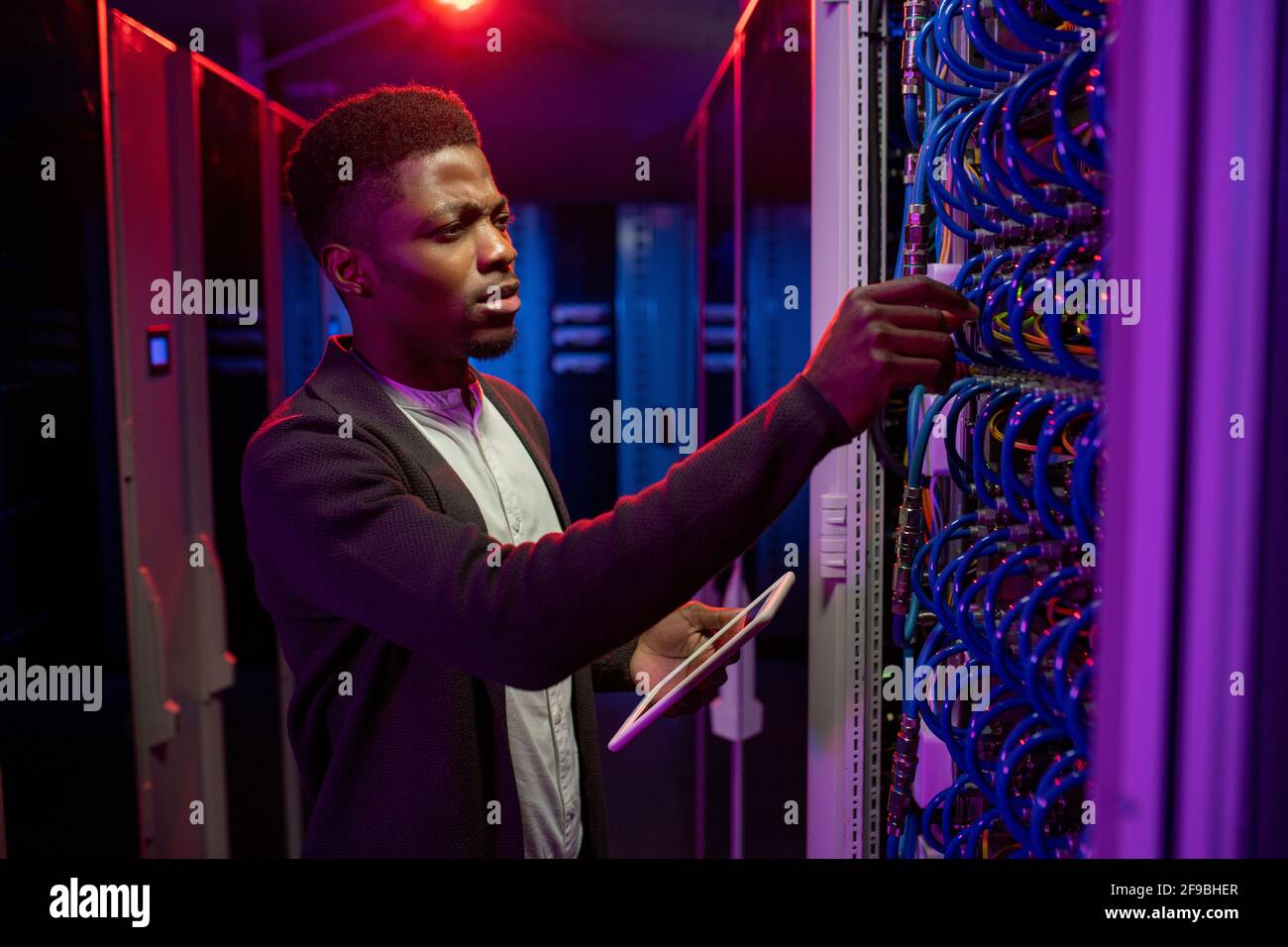 Concentrato giovane uomo nero utilizzando il tablet durante l'esame delle apparecchiature del data center in una stanza server buia Foto Stock