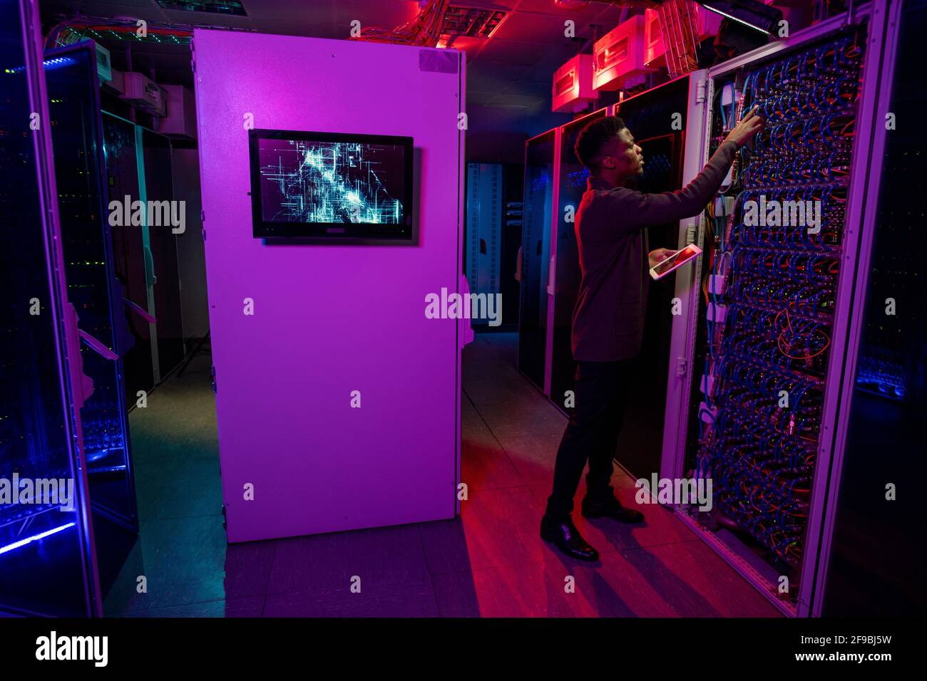 Serio giovane uomo nero in piedi con il tablet al cabinet del server e controllare il server blade durante la gestione dei server Foto Stock