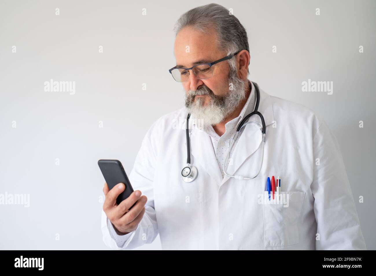 Ritratto di medico senior in camice bianco guardando il suo smartphone su sfondo bianco. Foto Stock