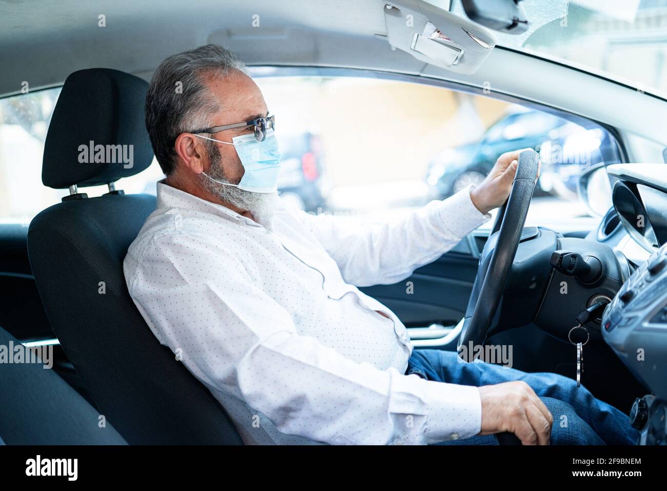 Un uomo anziano in una maschera medica che guida un'auto. Concetto pandemico di coronavirus. Viaggio su strada, viaggio e concetto di persone anziane Foto Stock