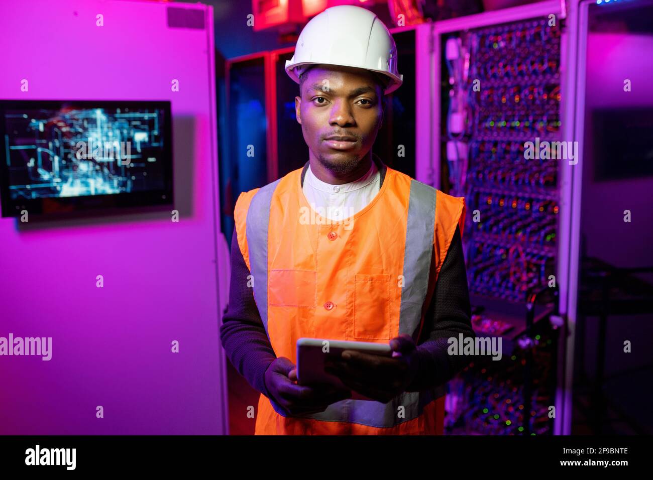 Ritratto di un serio e sicuro ingegnere di data center afro-americano giubbotto arancione e cappello bianco in piedi con tablet nel server camera con luce al neon Foto Stock