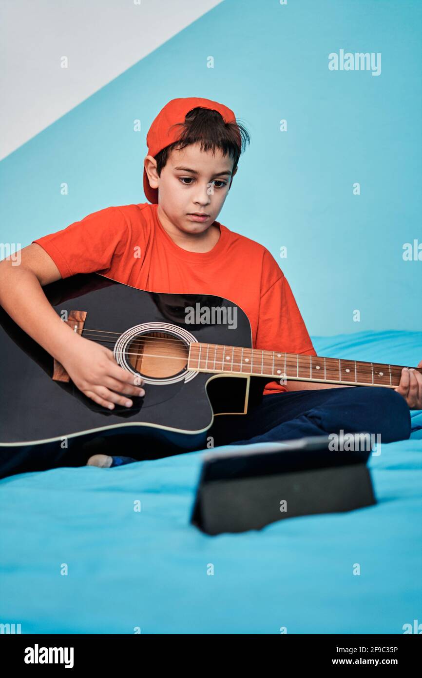 Ragazzo imparando a suonare la chitarra guardando il tablet sulla parte superiore di letto in camera Foto Stock