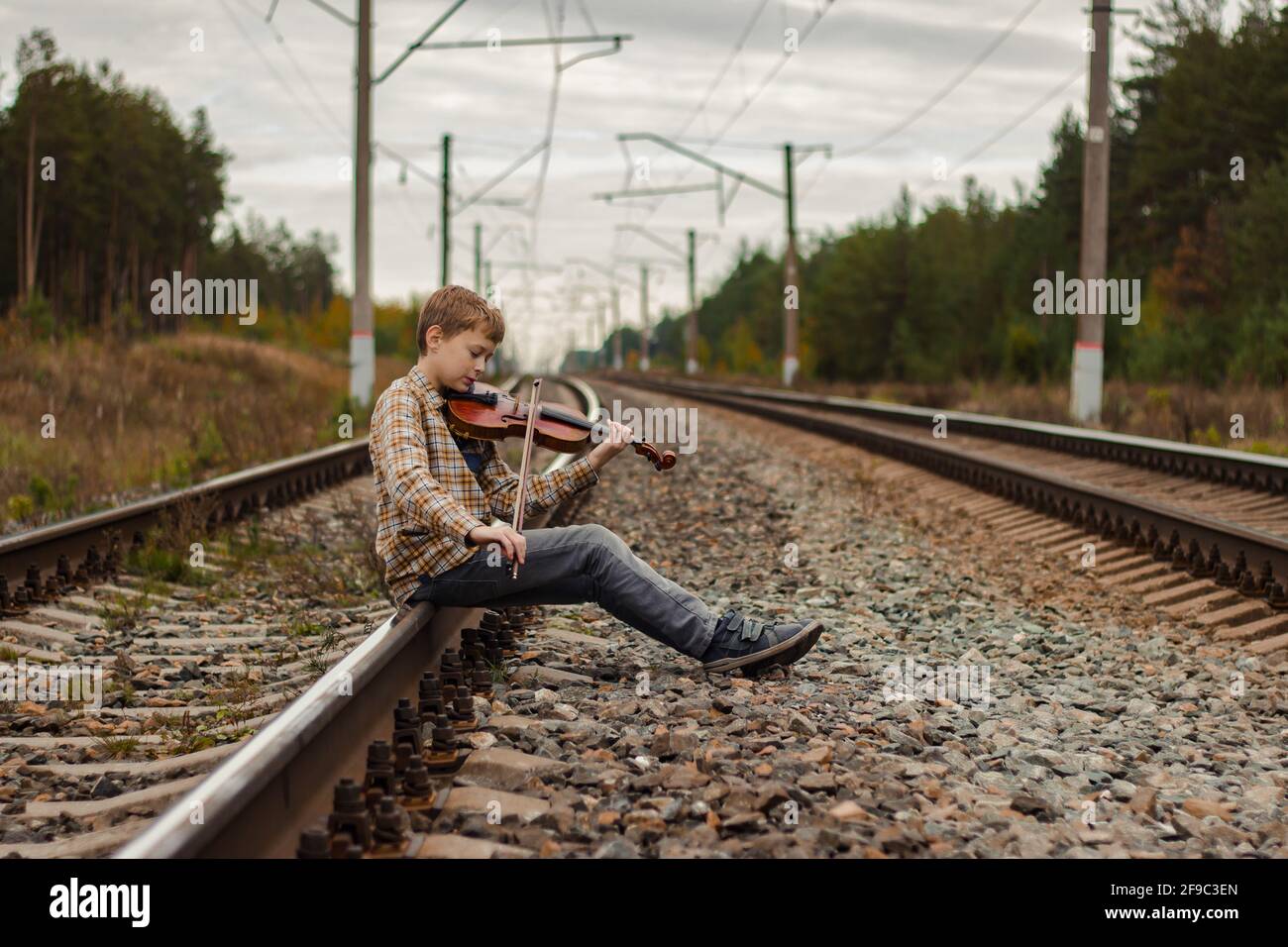 Un bel ragazzo biondo si siede sulle rotaie della ferrovia e suona il violino. Foto Stock