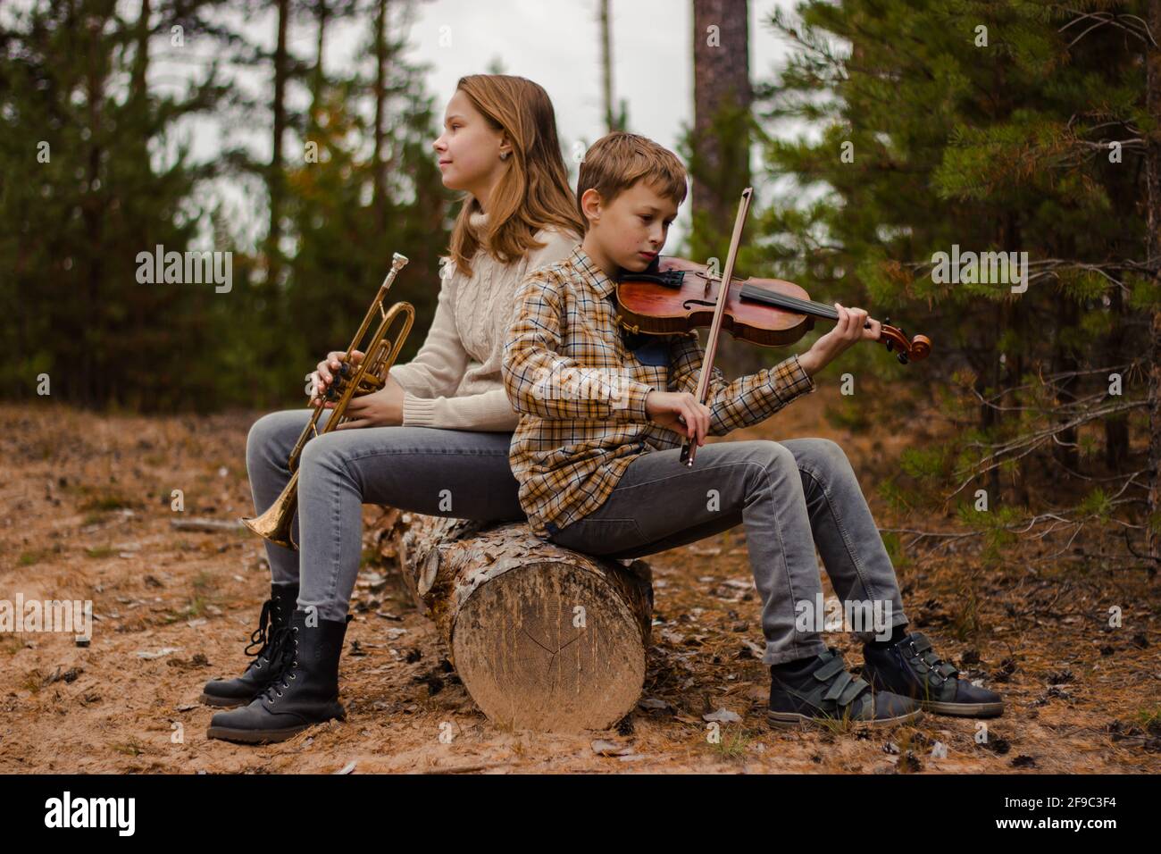 Ragazzo e ragazza, fratello e sorella, gli adolescenti suonano la tromba e il violino seduti nei boschi su un tronco. Foto Stock