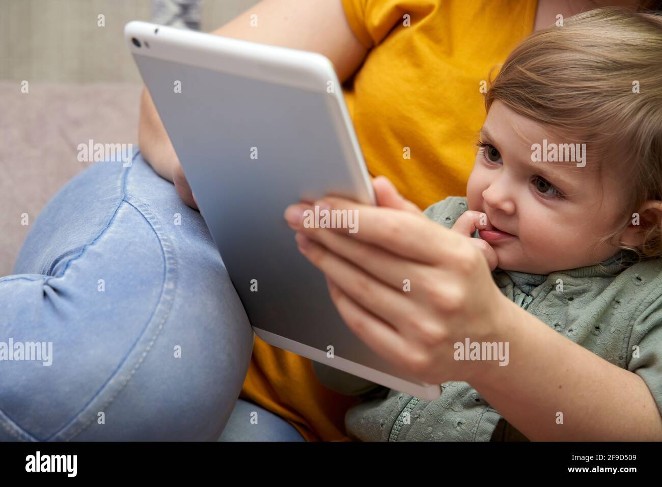 Un primo piano di una ragazza tra le braccia di sua madre che guarda insieme un tablet. Concetto di comfort e di svago a casa. Foto Stock