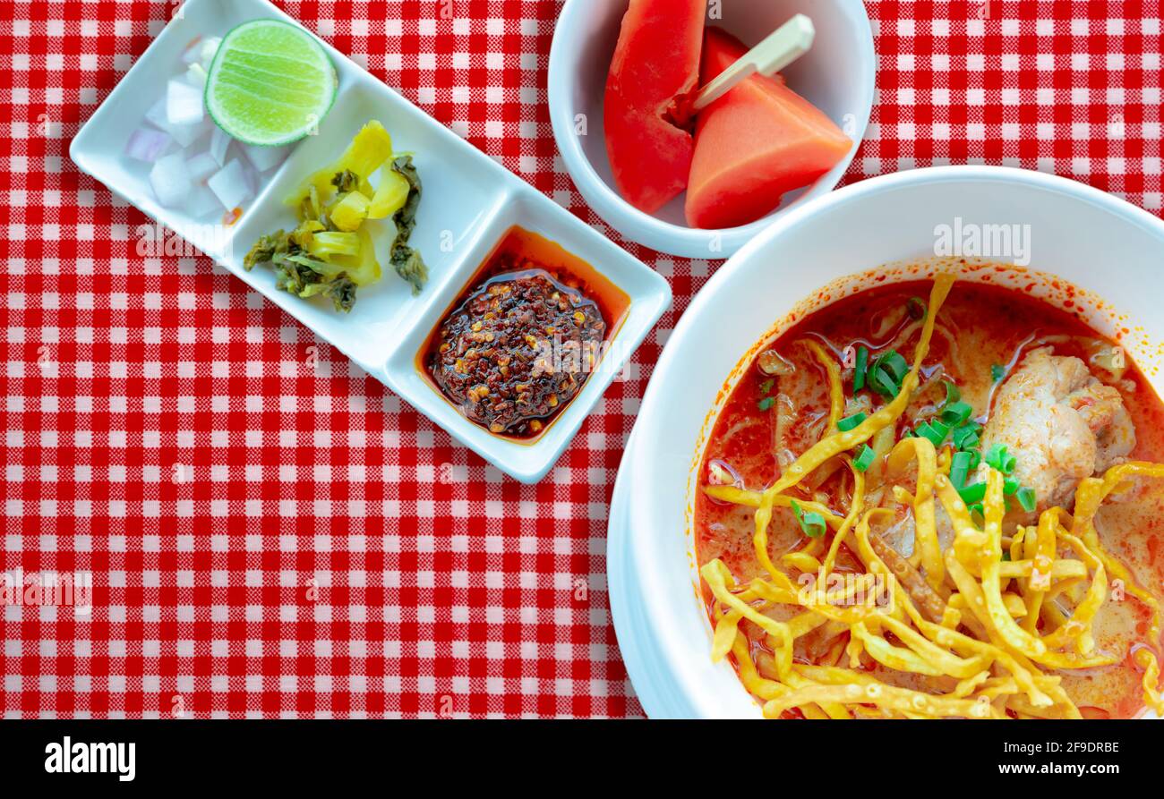 Zuppa di noodle al curry del nord della Thailandia con pollo e contorno in ciotola bianca su tovaglia a scacchi rossi. Vista dall'alto di Khao Soi e papaya matura con legno Foto Stock
