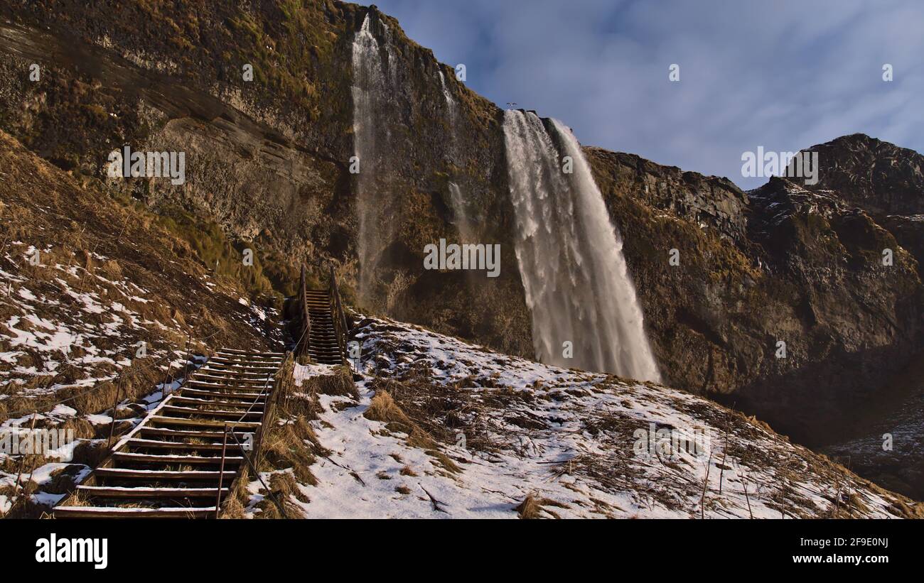 Splendida vista ad angolo basso della famosa cascata Seljalandsfoss sulla costa meridionale dell'Islanda, vicino alla circonvallazione, durante il sole giorno invernale con scale di legno. Foto Stock