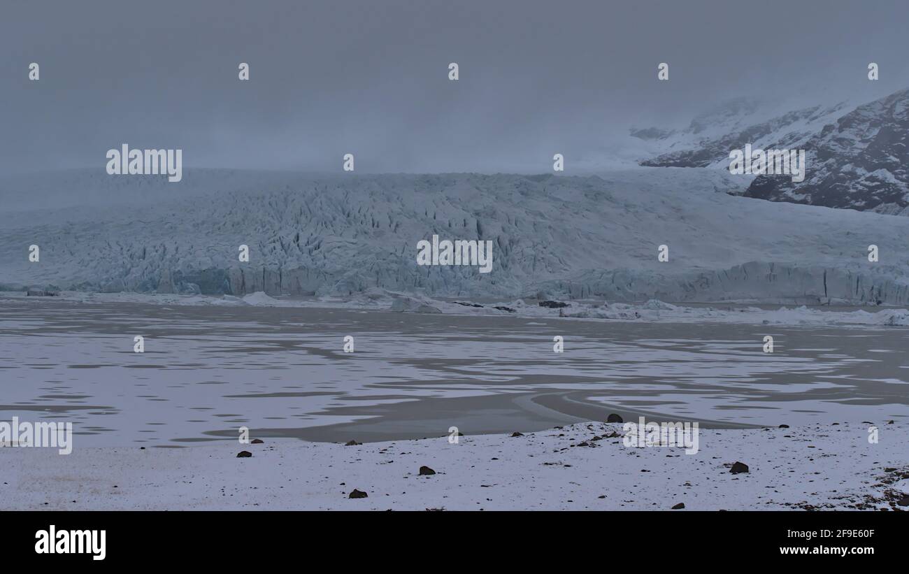 Vista frontale del lago glaciale di Fjallsárlón, nel sud dell'Islanda, con il ghiacciaio di Fjallsjökull, parte della calotta glaciale di Vatnajökull, e le aspre montagne tra le nuvole. Foto Stock