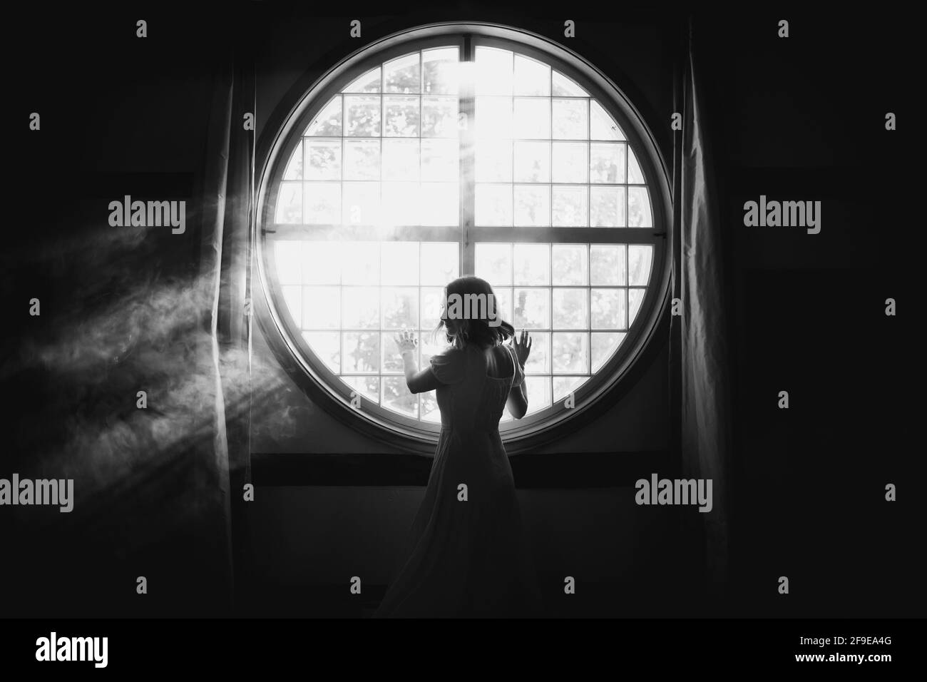Vista posteriore di una donna delicata irriconoscibile che guarda sopra la spalla recinzione a contatto su finestra rotonda in casa in luce solare Foto Stock