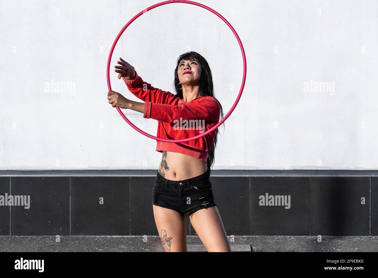 Giovane donna tatuata in abbigliamento sportivo che avvolge il cerchio di hula mentre ballano contro pareti di mattoni con ombre e guardando avanti alla luce del sole Foto Stock