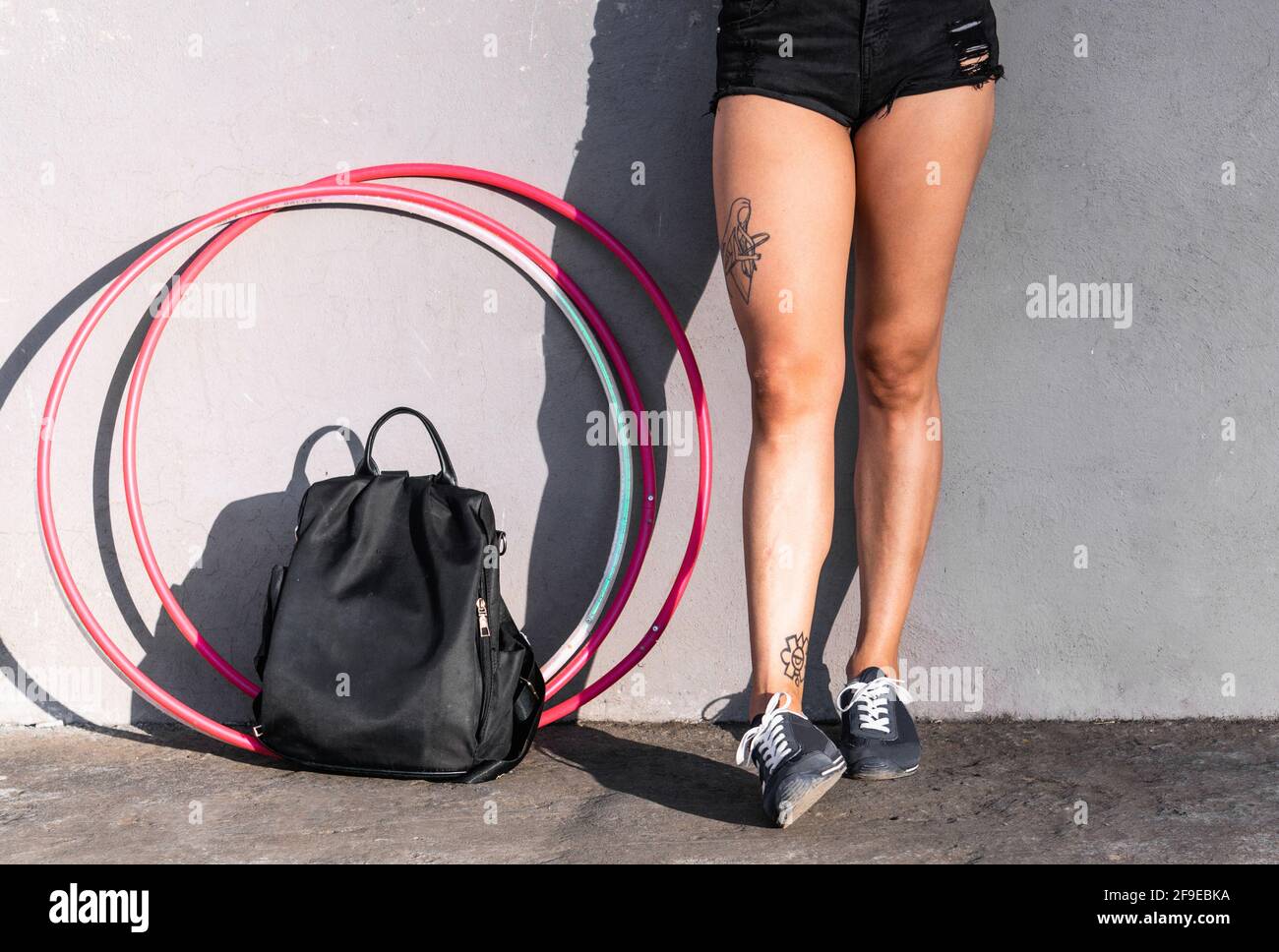 Sfaccia la donna irriconoscibile in pantaloncini corti e sneaker in piedi sul pavimento con zaino e cerchi in hula alla luce del sole su sfondo grigio Foto Stock