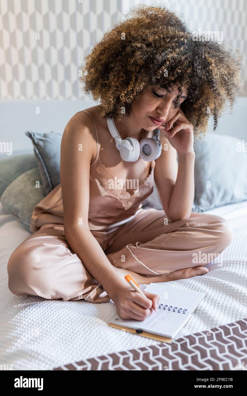 Giovane donna araba in abbigliamento da notte con capelli afro e wireless  cuffie che prendono appunti in blocco note mentre si tocca il viso nella  camera da letto Foto stock - Alamy