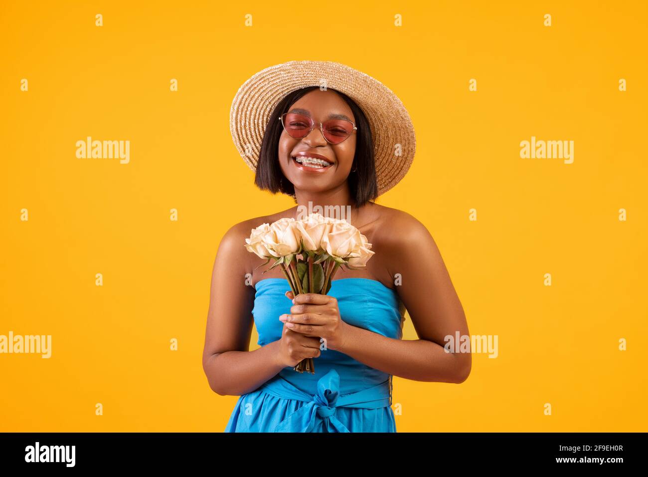 Bella donna nera in abito estivo e cappello di paglia tenuta bouquet di fiori su sfondo arancione studio Foto Stock