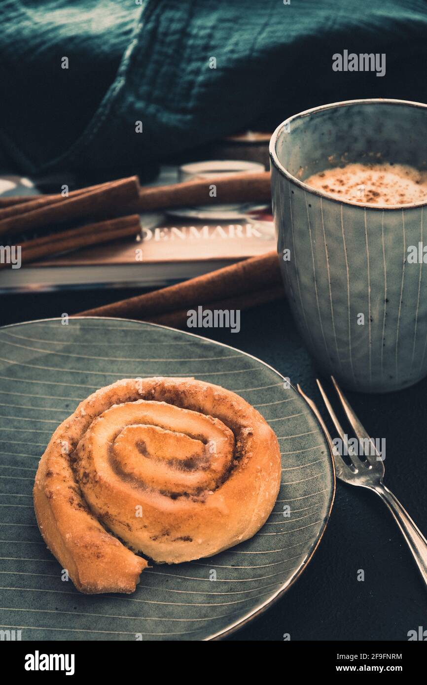Rotolo di cannella su un piatto grigio, caffè e forchetta. Sullo sfondo un libro, bastoni di cannella e un panno, colori di moody. Primo piano verticale. Foto Stock