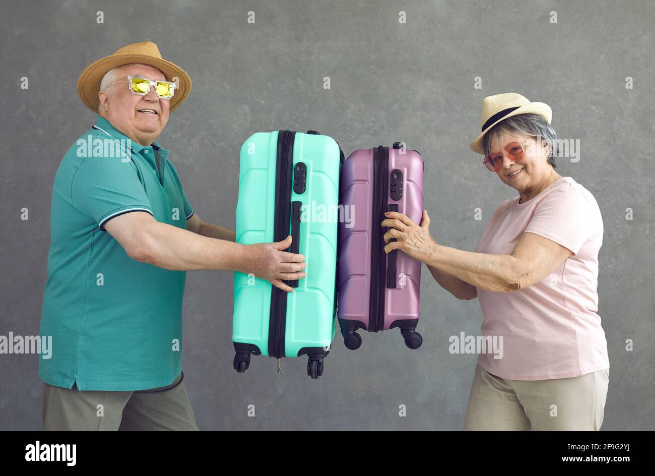 Divertente coppia sposata senior attiva di turisti con valigie luminose in mani su sfondo grigio. Foto Stock