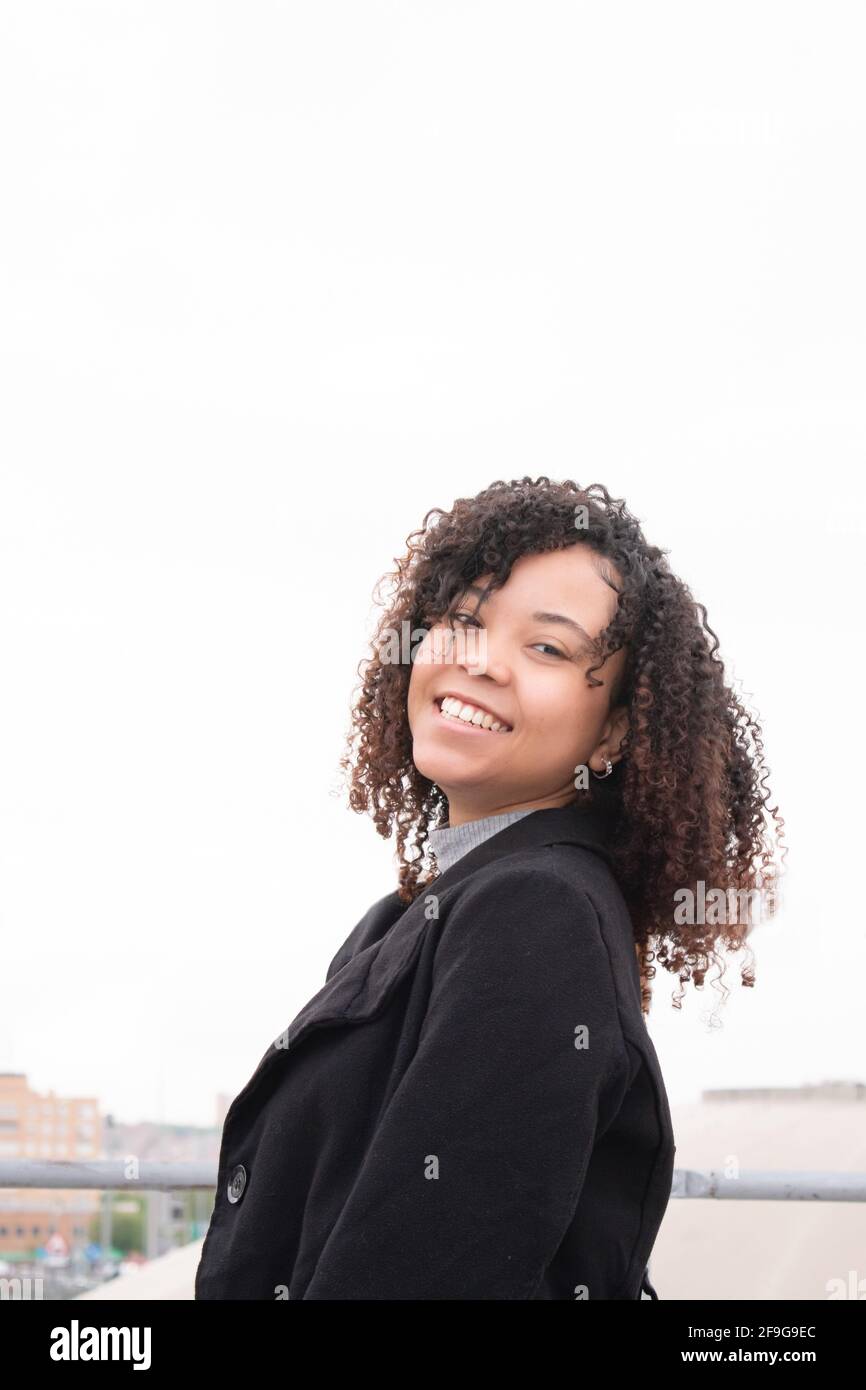 Donna nera sorridente con capelli ricci che guarda direttamente la fotocamera con il cielo di sfondo bianco. Foto Stock