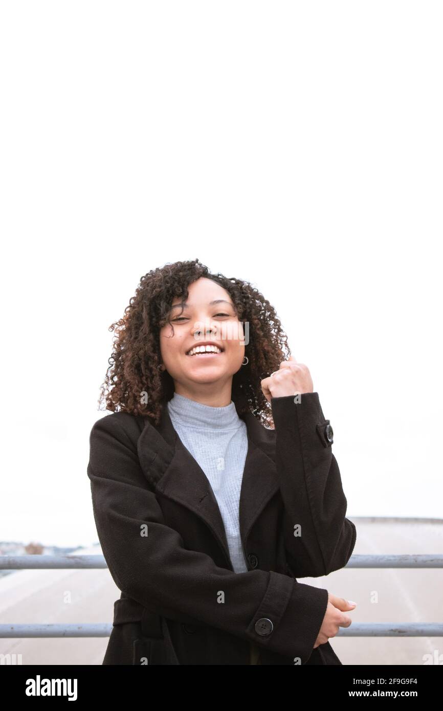 Giovane donna sorridente con capelli ricci che guarda direttamente la macchina fotografica con cielo di sfondo bianco vestito di lana nera. Foto Stock