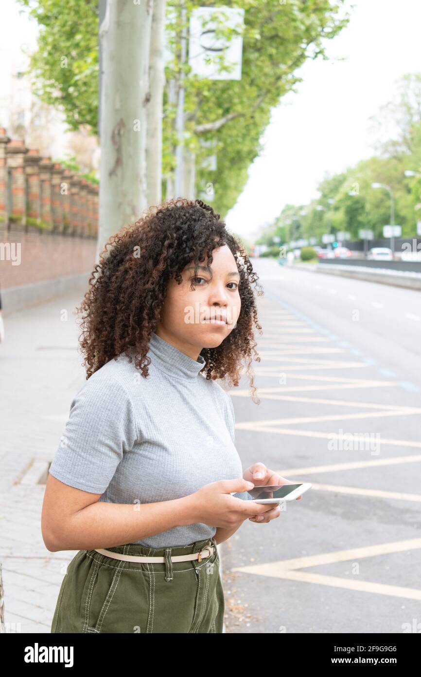 giovane ragazza con capelli ricci usando un telefono cellulare in attesa di un taxi o uber sulla strada. Foto verticale. Foto Stock