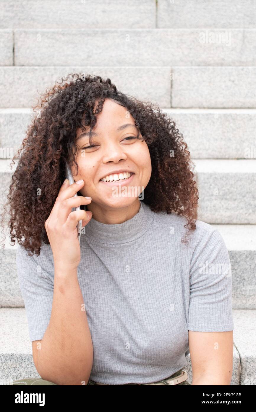 Giovane donna con capelli ricci che sorride mentre parla al telefono con le scale fuori fuoco sullo sfondo. Foto Stock