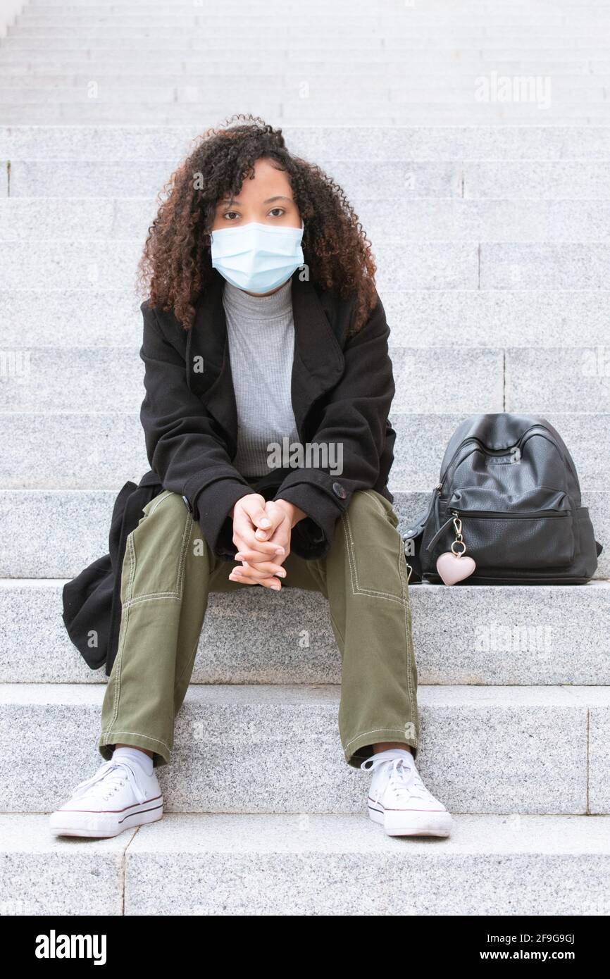 Giovane donna con maschera seduta sulle scale poggiando le braccia sulle ginocchia con le mani aggrappate guardando direttamente la fotocamera. Foto Stock