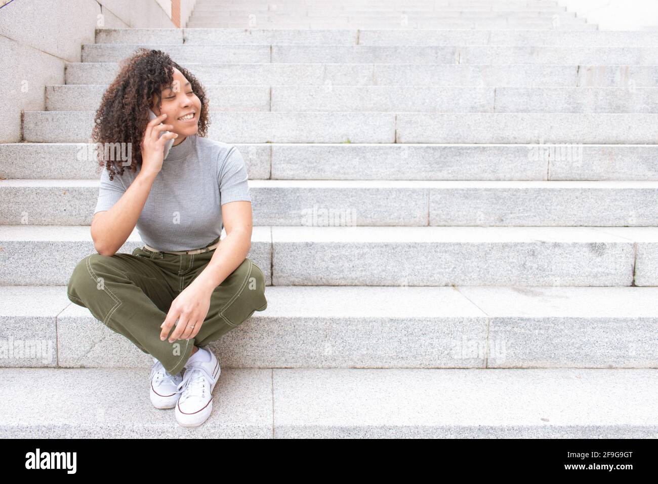 giovane donna nera con capelli ricci seduti su una scala parlando sul suo telefono cellulare mentre sorride e guarda a. il lato di uno spazio per la copia Foto Stock